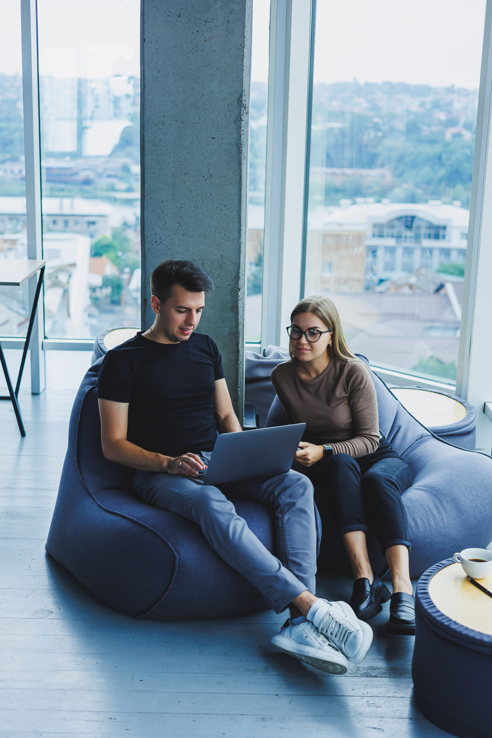 Students sit in the workspace near the window and work together on a project on a laptop. Cheerful colleagues managers look at laptop together and smile.