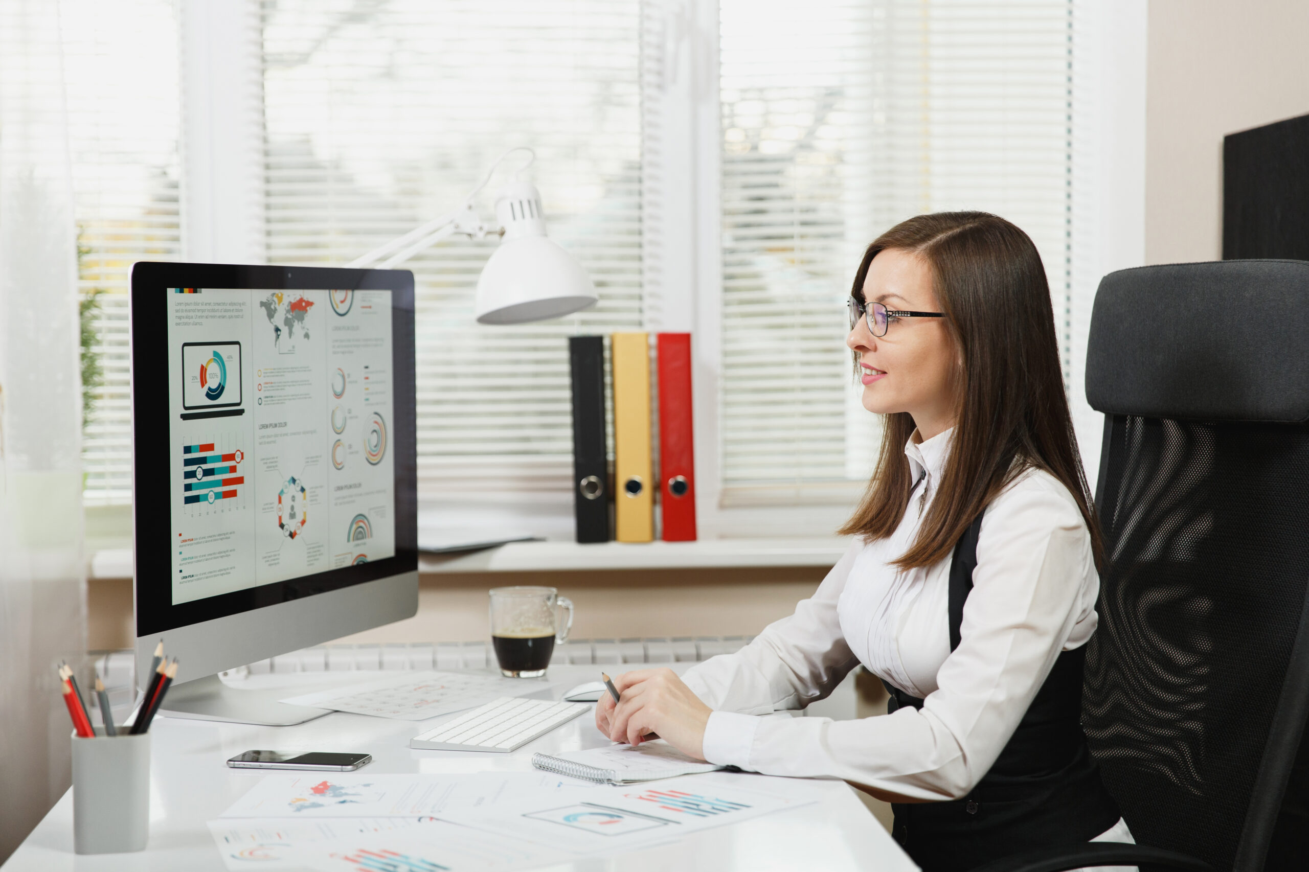 The beautiful smiling brown-hair business woman in suit and glasses sitting at the desk, working at computer with documents in light office, looking at the monitor